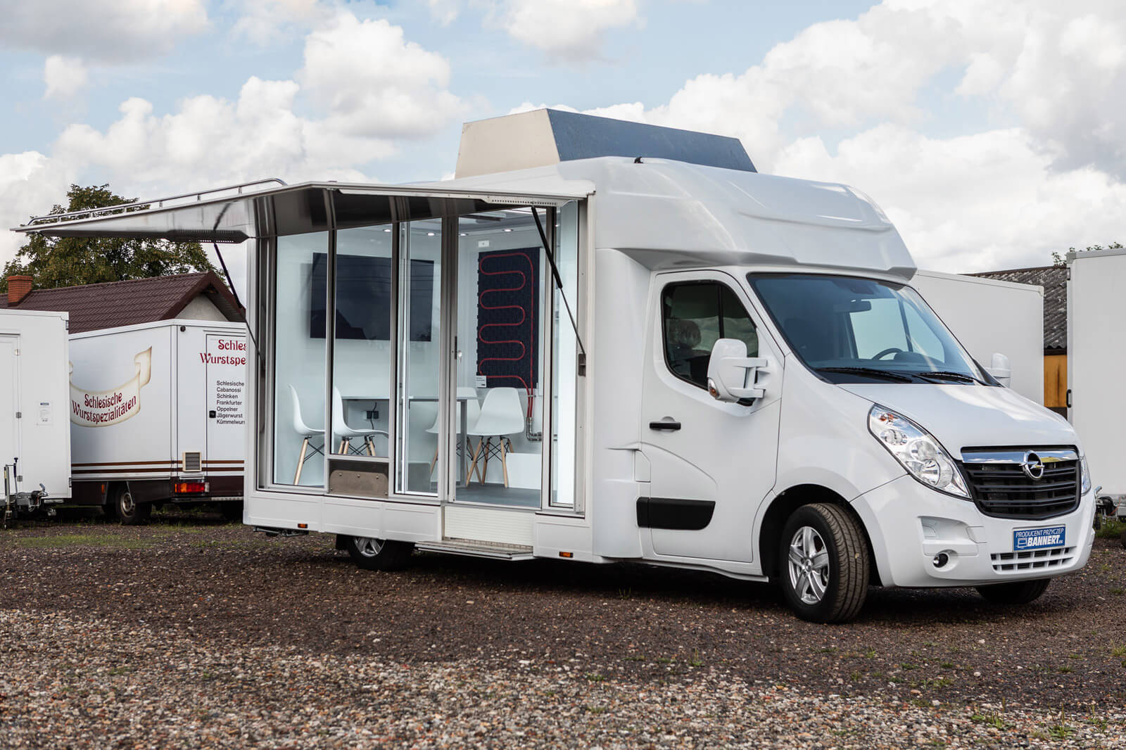 Biały van z przeszklonym bokiem, służący jako mobilne biuro lub showroom z krzesłami i ekranem w środku.